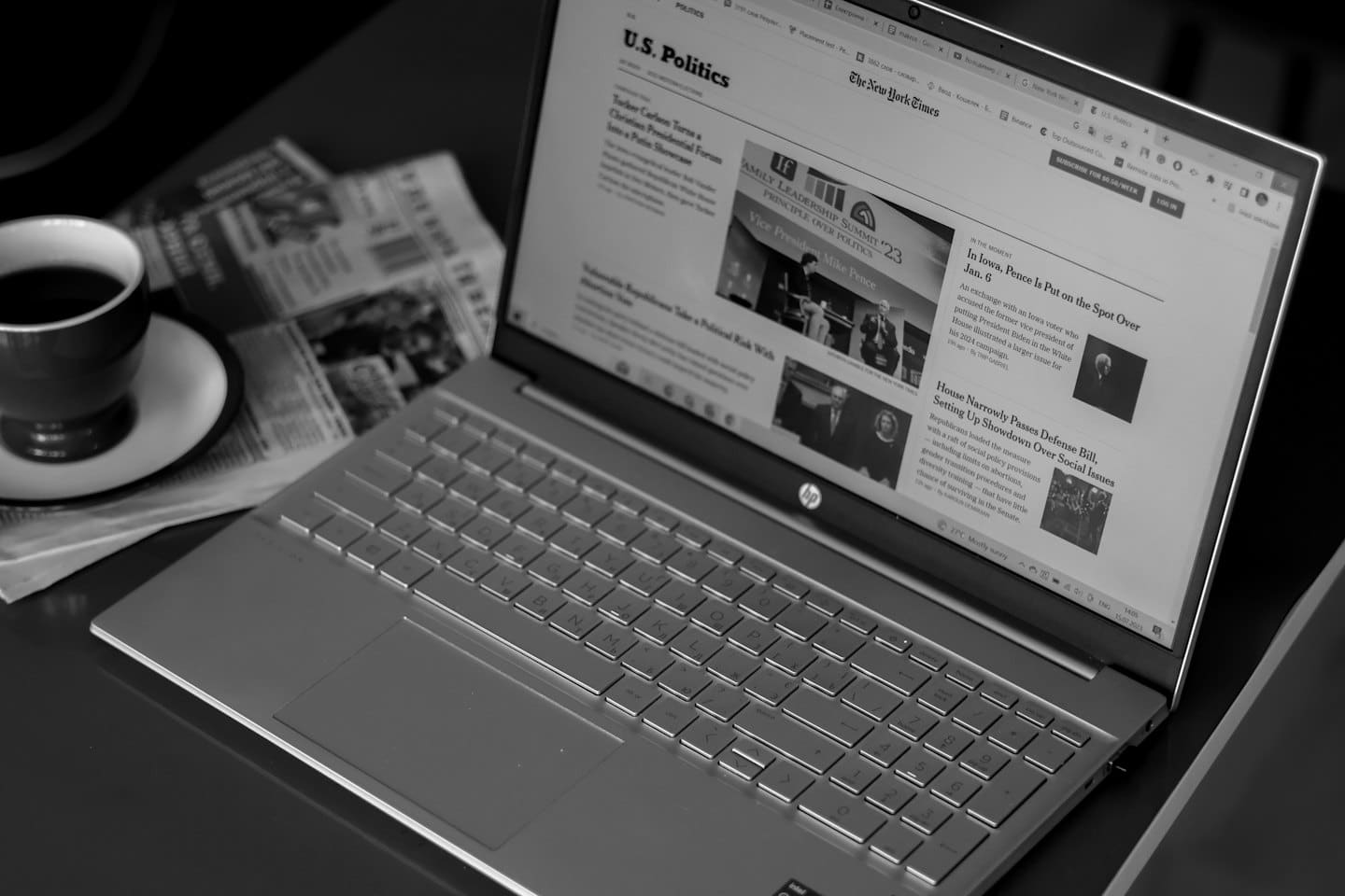 Portátil mostrando artículos de noticias con café y periódicos sobre el escritorio.